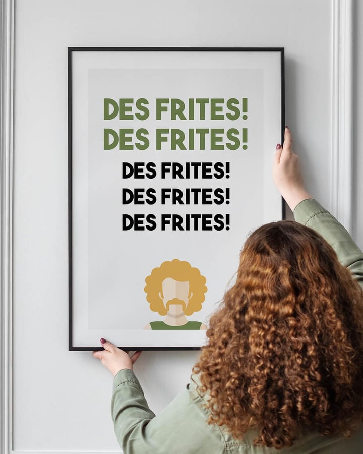 Une femme aux cheveux bouclés accroche l’affiche Des frites, dans un cadre noir, sur un mur blanc à moulures