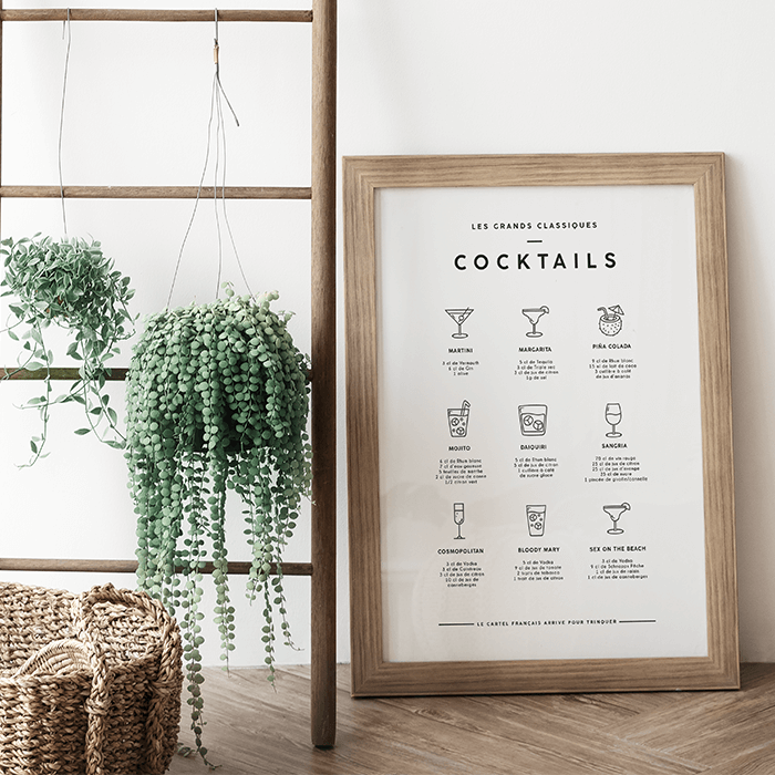 L’affiche Cocktails dans un cadre en bois clair, posée sur un parquet à côté d’une échelle et de plantes suspendues
