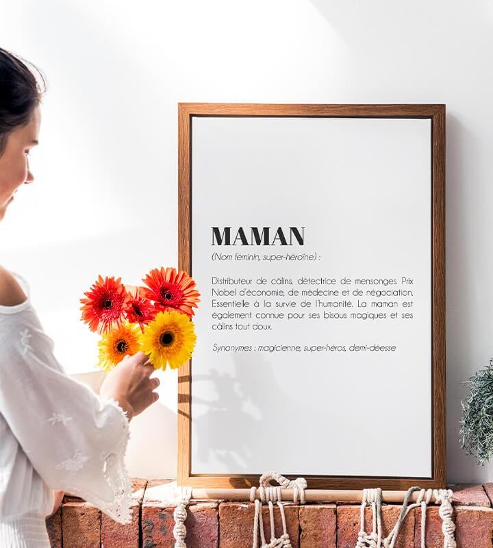 Une femme tient un bouquet de gerberas près de l’affiche Définition Maman, posée dans un cadre en bois