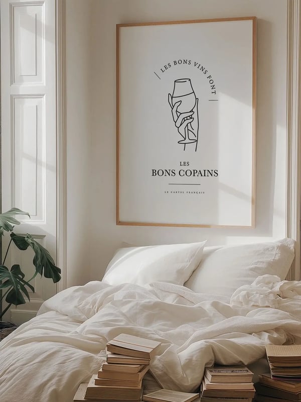 Affiche Les bons vins font les bons copains dans un cadre en bois clair, au-dessus d’un lit aux draps blancs