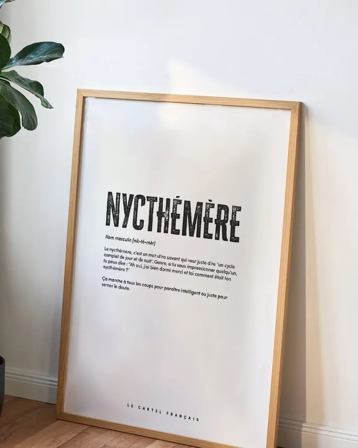L’affiche Nycthémère dans un cadre en bois clair, posée au sol contre un mur blanc, à côté d’une plante