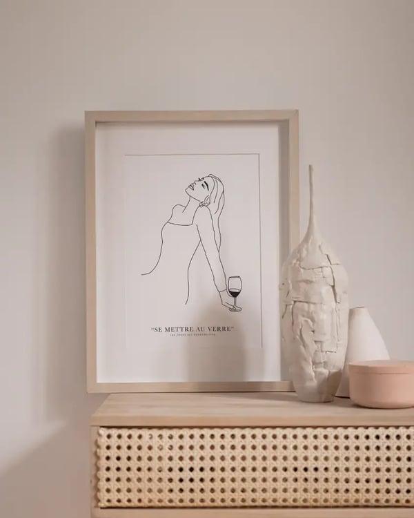 Affiche Se mettre au verre dans un cadre en bois clair, posée sur une console en rotin à côté de vases