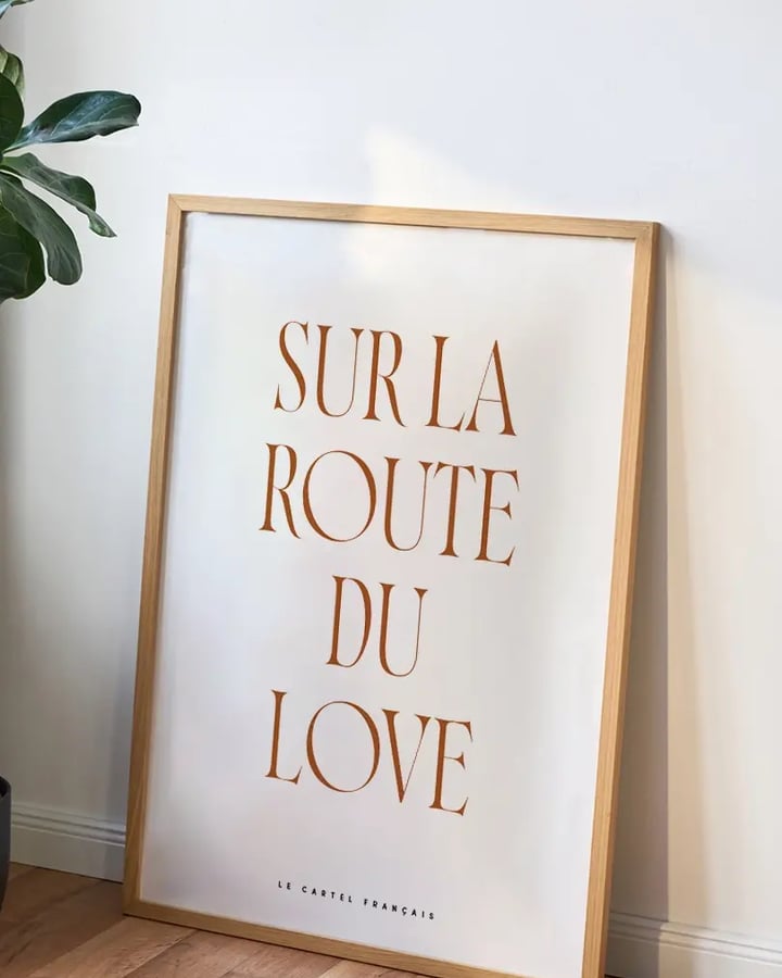 L’affiche Sur la route du love dans un cadre en bois clair, posée sur un parquet contre un mur blanc