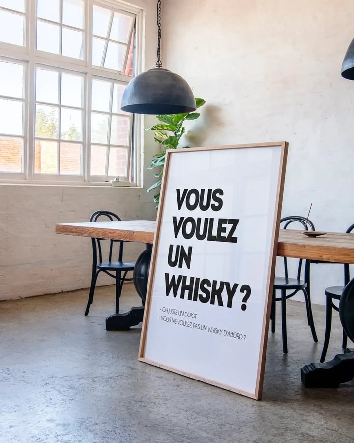 L’affiche Vous voulez un whisky dans un cadre en bois clair, posée au sol près d’une grande table