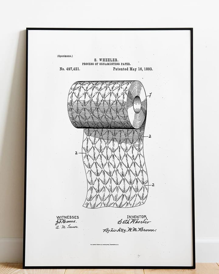 L’affiche Brevet papier toilette dans un cadre noir, posée sur un parquet clair contre un mur blanc